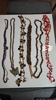 Photo shows six costume jewelry necklaces arranged side by side on a white surface.
