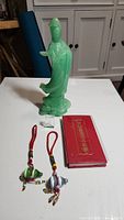 Green Asian-style figurine on clear base, red book, two fish charms with red cords, and small clear cube displayed on white surface.
