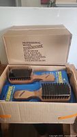 Open cardboard box displaying six Supergroomer Club Brushes inside, showing natural wood handles and dark boar bristles. The box label indicates 100% natural boar bristles and durable wood pallet.