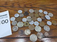 Top-down view of 36 assorted international coins laid out on a wooden surface, showing variety in size, color, and design including one coin with a hole in the center.