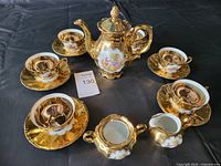 Full tea set arranged on black background showing the teapot, four cups and saucers, sugar bowl, and creamer, all with reflective gold finish and painted scenes.