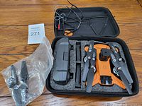 Photo of opened carrying case displaying foldable orange drone with black propellers, extra propellers in plastic bag, and accessories in foam cutouts.