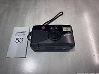 Camera front view showing body, lens cover closed, and wrist strap attached