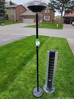 Photo showing a black floor lamp with basket-style shade and a silver tower fan placed on grass outdoors.