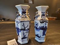 Front and side views of two matching vintage porcelain vases with blue painted figures and floral designs on white hexagonal bodies. Wide flared square rims and double ring handles visible on necks.