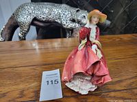 Front view of the Royal Doulton figurine showing the woman in a red dress and orange hat on a wooden surface, with a silver leopard sculpture in the background and a numbered auction tag (115) nearby.
