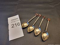 Four silver tone decorative spoons with reddish-brown inset tops laid side by side on a black surface with a lot number card