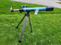 Full view of the blue Sky-Watcher telescope mounted on an adjustable tripod standing on grass with a road in the background.