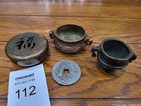Four vintage Asian decorative items displayed on wooden surface: two small bronze vessels with handles, one small round bronze lid with Asian character cutout, and one large circular metal coin with square hole in center.