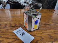 Front view of the square enamel holder with detailed painted Asian figures and metal handle, alongside the metal tea pot.
