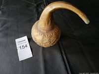 Vintage brass water dipper on black surface showing front side detail with embossed designs and long curved spout.