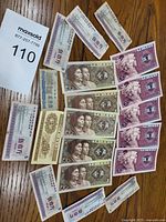 Image showing a collection of 13 vintage Chinese paper currency notes arranged on a wooden surface. The notes depict various denominations, with predominant colors including purple, brown, and beige. Included is a bidding card showing the lot number 110.
