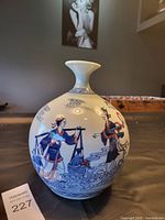 Front view of round white porcelain vase with blue painted Asian figures carrying items, placed on table with dark background.