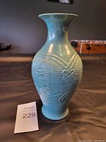 Front view of turquoise blue ceramic vase with raised swirl and geometric design on body, standing on flat surface next to Maxsold number card 229.