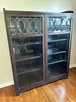 Front view of antique wooden display cabinet showing two glass doors with upper decorative leaded glass panels featuring circular patterns. Solid dark wood frame and shelves visible inside.