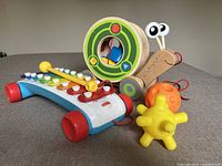 Wooden snail toy with green spiral shell and colorful wooden blocks inside, colorful xylophone with yellow mallet, and two sensory balls in yellow and orange on gray surface.