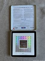 Opened tin box with internal metallic lid displaying bilingual text about the commemorative coin project and holographic coin set holder with '1999' year in center.