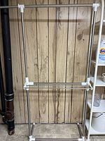 Full view of the metal clothes drying rack standing against wood panel wall, showing the horizontal bars and plastic joints.