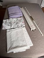 Photo showing three folded curtains (white, purple floral, white lace) laid out on bed with white window blind and white tension rod with clamps