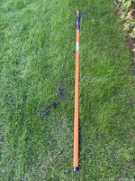 Full length view of the Fiskars telescoping manual tree pruner laid on grass, showing orange pole, black handle, and attached rope pull.