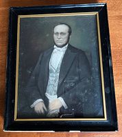 Full view of the framed portrait showing the man seated wearing dark formal clothing and framed in a black wooden frame with gold inner trim.