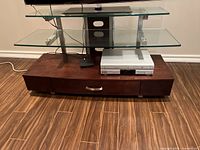 Front view of the glass top and middle shelf with electronic devices on the wooden base showing the central drawer and compartments.