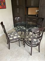 Five-piece dining set with round glass top table and four upholstered chairs arranged on tiled floor in front of dark wood cabinetry.