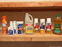 Photo showing nine assorted household cleaning products arranged on a wooden shelf, including spray bottles, plastic jugs, and small bottles of various sizes and brands.