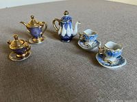 Miniature tea set items arranged on fabric surface showing teapot, coffee pot, sugar bowl and two cups with saucers