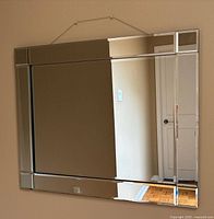 Front view of rectangular wall mirror showing reflective glass frame and hanging chain; mirror reflects a room with door and floors.