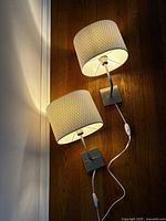 Pair of IKEA sconce lamps illuminated, showing lampshades and curved arms with square base plates and attached power cords.