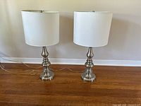 Pair of matching table lamps with silver metal bases and beige fabric drum shades placed on a hardwood floor against a wall, lamps powered off.