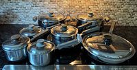 Full set of Lagostina stainless steel pots and pans with lids arranged on stovetop reflecting light and kitchen backsplash.