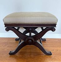 Side profile showing dark wood X-style curved legs and upholstered beige cushion with nailhead trim.