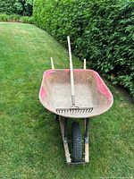 Front view of heavily used red metal wheelbarrow with wooden handles and inflated black tire with metal garden rake lying inside.
