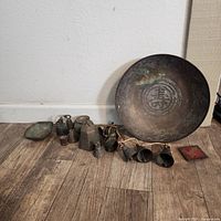 Photo shows the full set of various sized metal bells, a large round ornate dish or gong, and a decorated small tray all placed on wooden floor.
