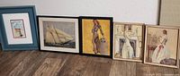 Six framed artworks arranged on floor against wall, includes miniature, sailing ship, pin-up, and three vintage women prints