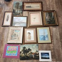 Lot of 11 framed pictures arranged on the floor showing diverse styles including landscapes, portraits, colorful abstracts, and nautical scenes.