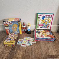 Photo showing Simpsons 1000-piece puzzle box, Simpsons super-size dominoes with cards spread out, Duff beer can figurine with Homer Simpson figurine on top, and a stack of Simpsons books.