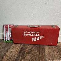 Red metal Milwaukee Sawzall case with handle and latches resting on floor next to sealed saw blade pack.