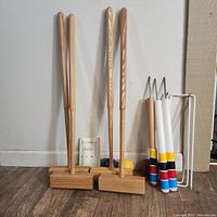 Full view of wooden croquet mallets, croquet balls in multiple colors, white hoops and metal stake against a white wall and wooden floor.