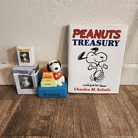Photo showing the Peanuts Treasury book by Charles M. Schulz next to a Snoopy music box featuring Snoopy on colorful stacked books labeled with school subjects, and two boxed Peanuts Snoopy ornaments.