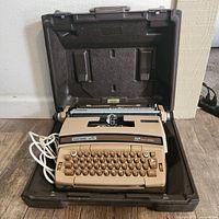 Side angled view of Smith Corona Coronamatic typewriter in open brown hard case on floor showing keyboard, paper roller and power cord.