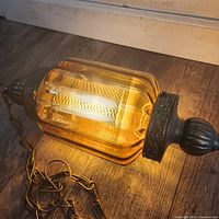 Amber glass hanging lamp lying horizontally on wooden floor with attached metal chain. Amber glass body with decorative metal caps and finials.