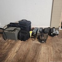 Photo shows six vintage camcorders arranged on floor alongside three camera carrying cases, showcasing exterior conditions and general size.