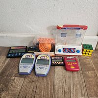 Photo showing entire lot of vintage electronic handheld games and small toys arranged on a floor against white wall