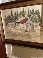 Framed watercolour painting showing rustic wooden buildings with evergreen trees in the background, signed by the artist.