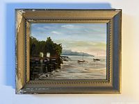 Frontal view of the framed antique oil painting showing the harbor scene with boats and dock under ambient light.