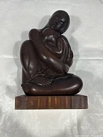 Side view of dark brown wooden sculpture depicting seated adult figure holding infant, on rectangular wooden base.