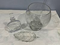 Clear glass punch bowl with ladle, rectangular decanter with stopper, and leaf-shaped serving plate on white fabric background